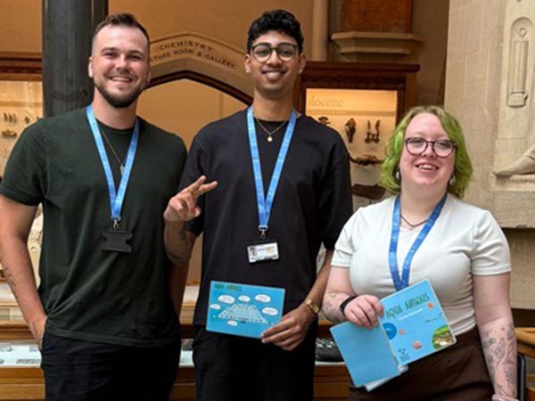 Three people standing indoors in a museum gallery, wearing name badges and smiling at the camera; display cases and exhibits are visible behind them.