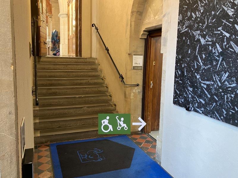 Interior view of a stone stairway inside the museum with a handrail and a green sign showing wheelchair and stroller symbols pointing right toward an accessible route.