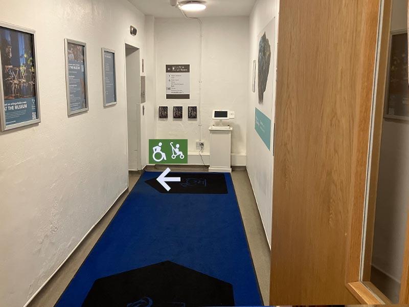 White-walled museum hallway with blue carpet runner and wall signage for visitor facilities, with a green wheelchair and stroller symbol sign pointing left toward the accessible route.