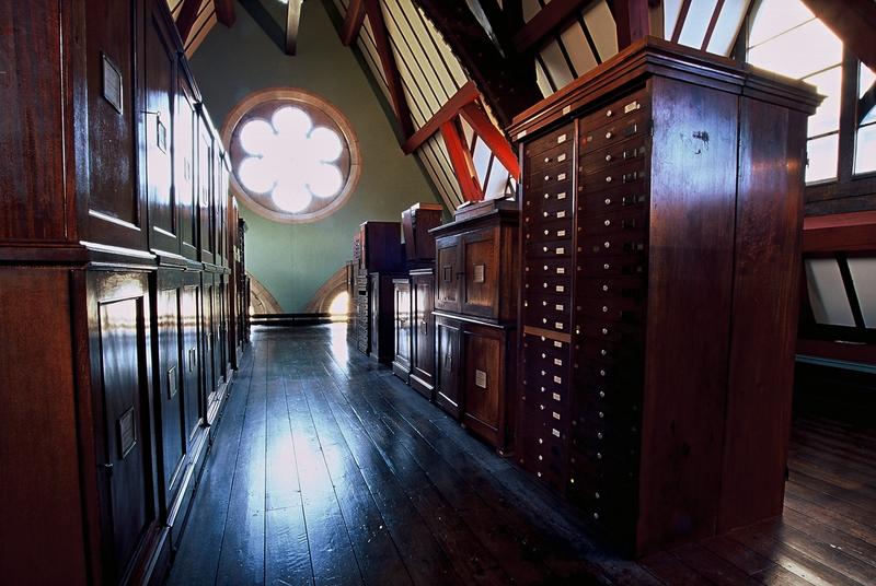Photograph of wooden-beamed and wooden-floored room with bright light emerging through flow-shaped window in back hall. In the room are dark wooden museum cabinets of drawers.