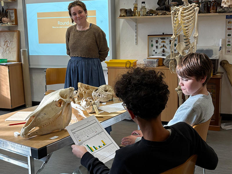 An educator stands beside a table of animal skulls while two young people review a worksheet in a classroom with a skeleton model in the background