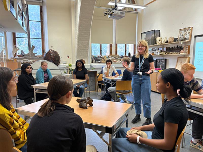 A teacher stands and gestures while speaking to a group of students seated in a classroom, with natural history specimens and large windows visible behind them.