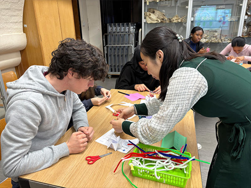 A student sits at a table while another person leans over to help them assemble a small craft using paper, scissors, and colourful pipe cleaners, with more students working in the background.