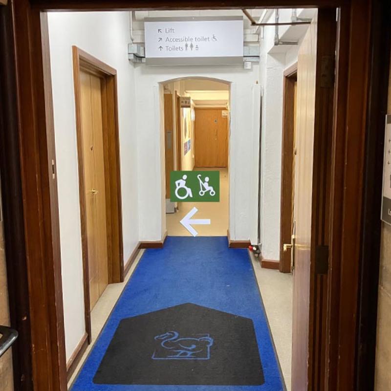 Museum corridor with wooden doors and overhead signage directing to the lift, accessible toilet, and toilets, with a green wheelchair and stroller symbol sign pointing left