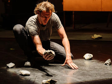 Performer crouching on a dimly lit stage, holding a piece of stone surrounded by scattered rock fragments.