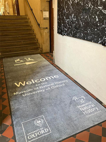 Accessible entrance to the Museum of Natural History, University of Oxford. A grey floor mat welcomes visitors and displays the museum and university logos, with an accessibility symbol and arrow pointing towards a lift on the right..