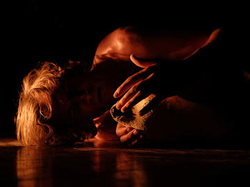 Performer lying on the floor in warm, low lighting, holding a piece of stone close to their face.