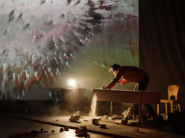 Performer leaning over a table as dust pours to the floor, with abstract imagery projected across the backdrop.