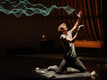 Performer kneeling onstage, holding up a piece of stone beneath a projected blue wave pattern.