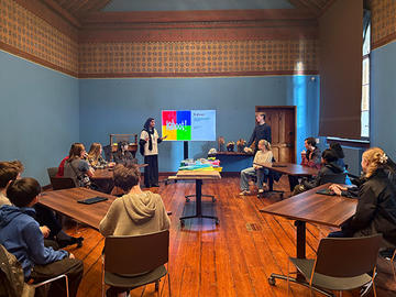 Students sit around tables in a classroom while two presenters stand at the front beside a screen displaying a colorful slide. Specimens and materials are arranged on tables in the centre.