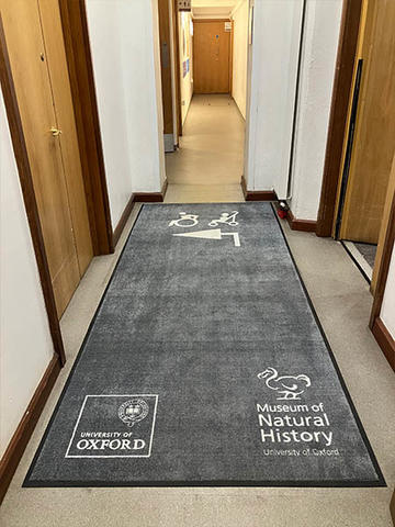 Grey floor mat displaying the Museum of Natural History and University of Oxford logos, with an accessibility symbol and arrow directing visitors towards the lift on the left.